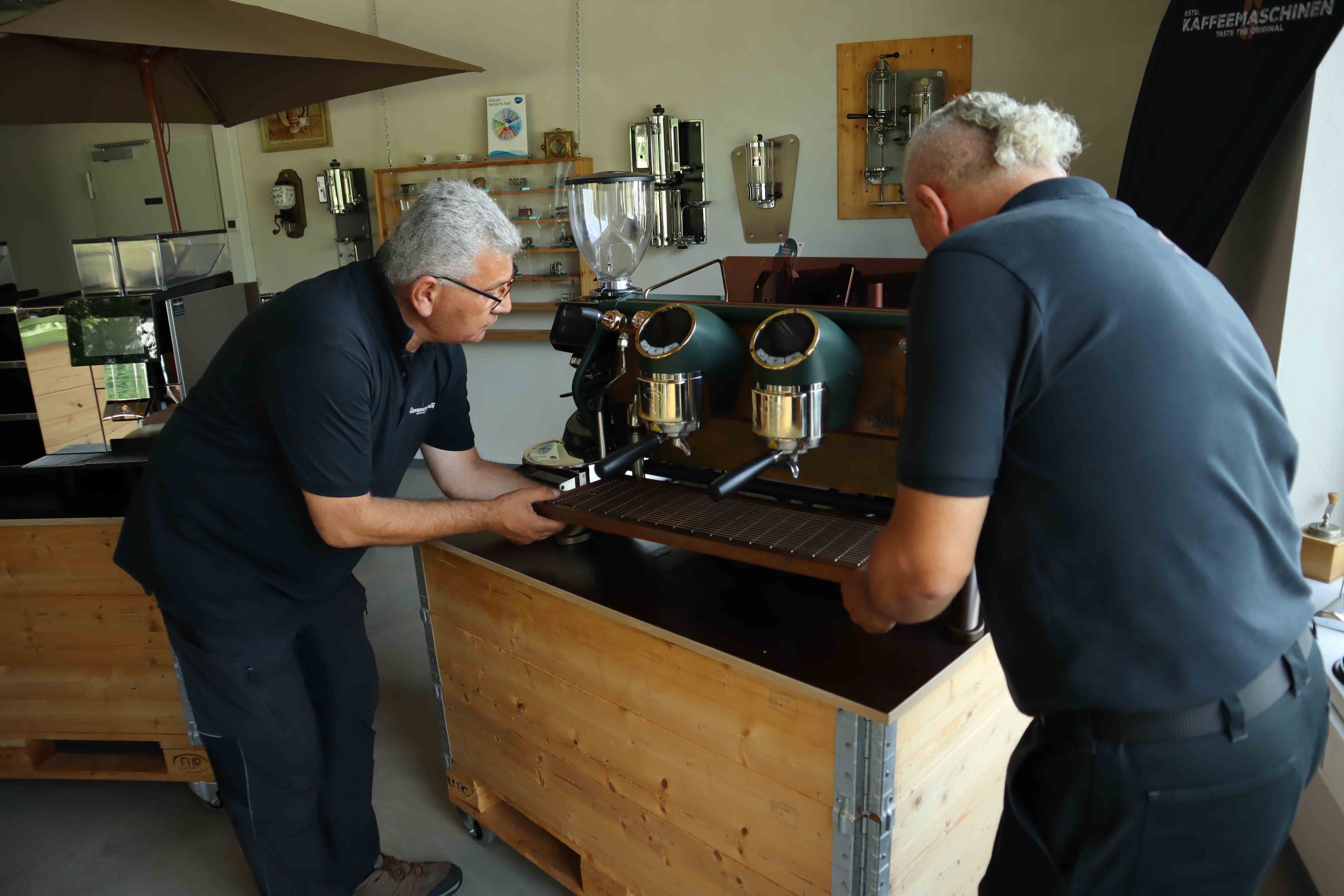 Two men in black shirts installing or adjusting a large espresso machine on a wooden counter indoors.