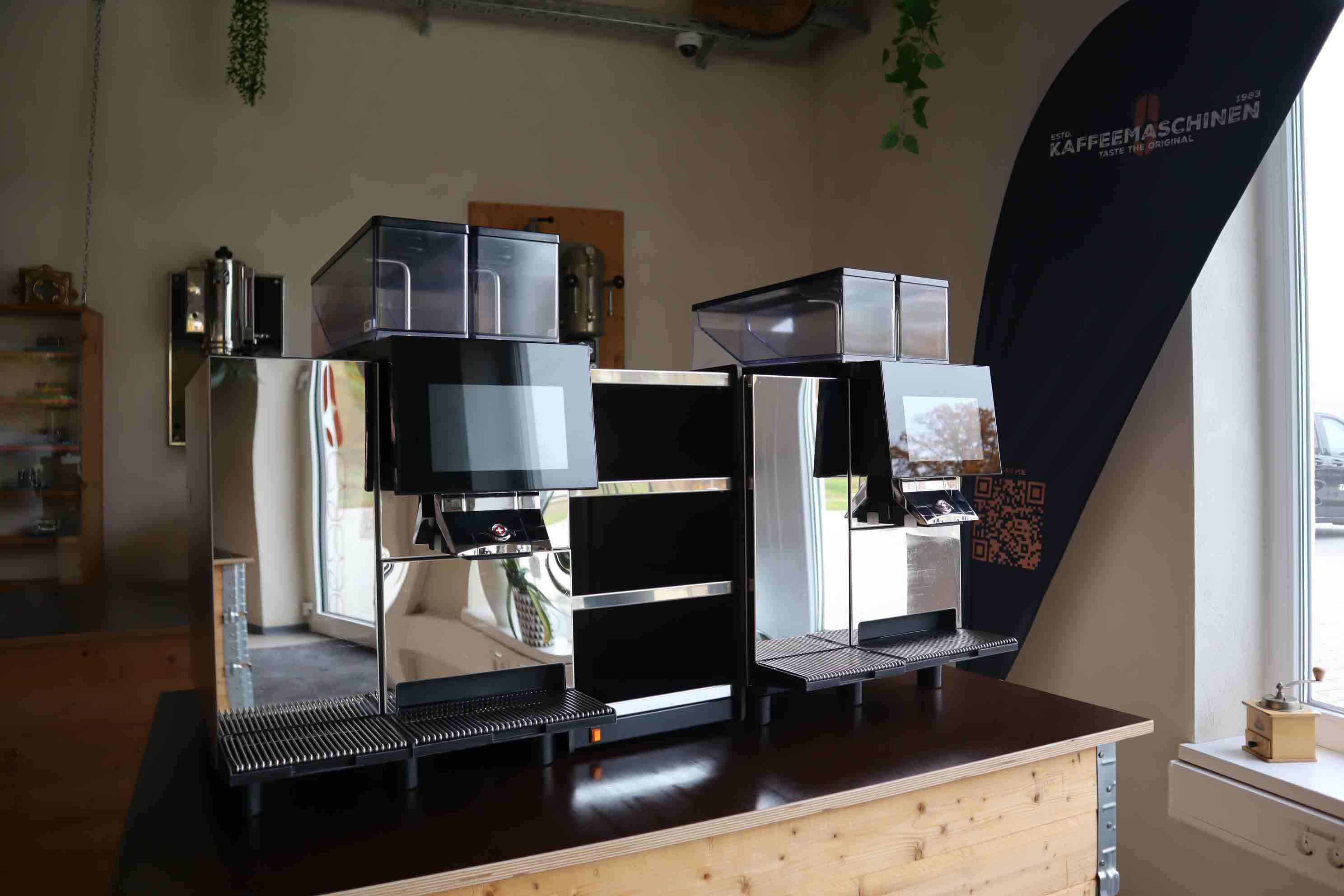 Two modern stainless steel coffee machines with digital displays on a wooden counter near a window.