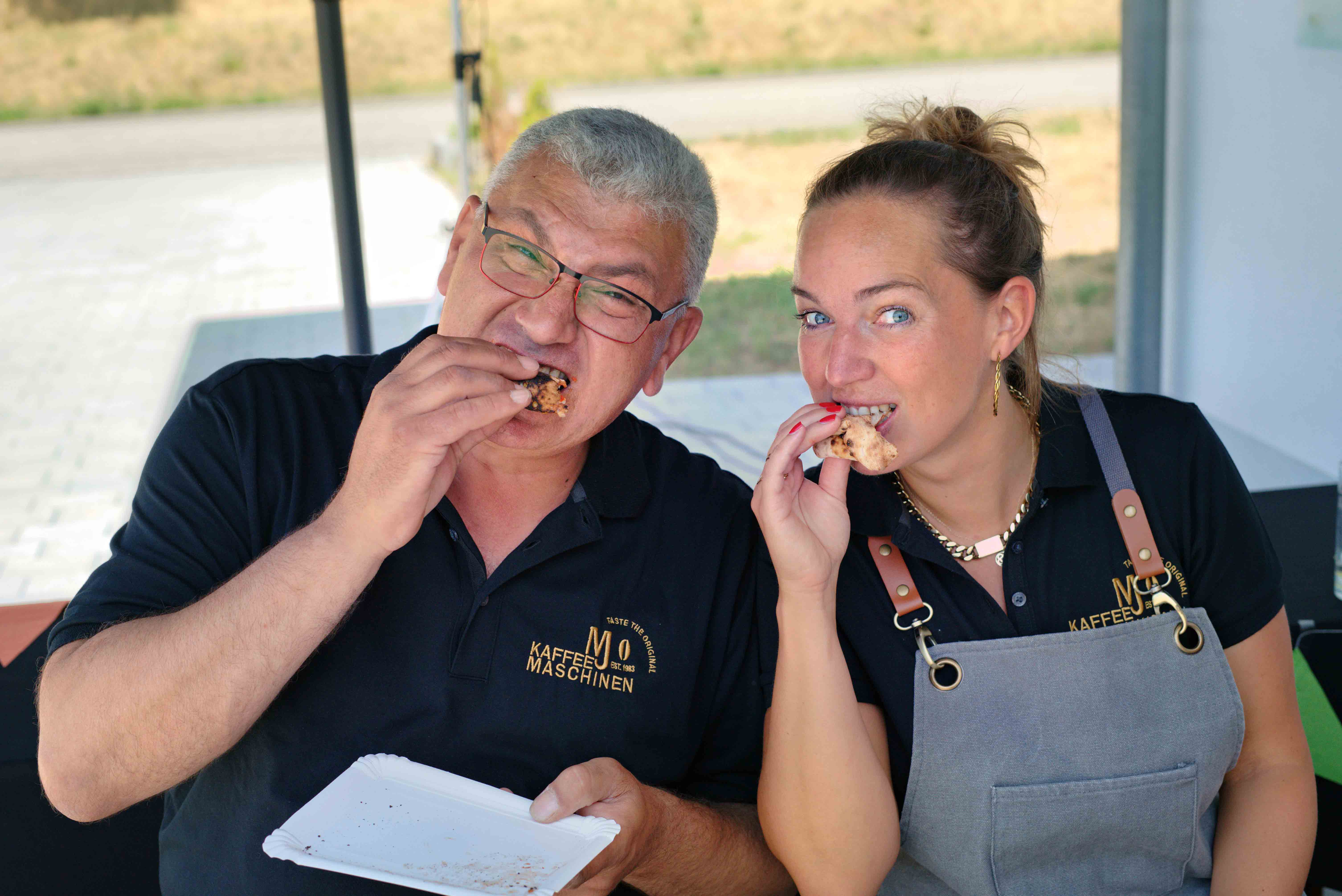 Two people wearing black shirts with company logos eating pizza slices outdoors.