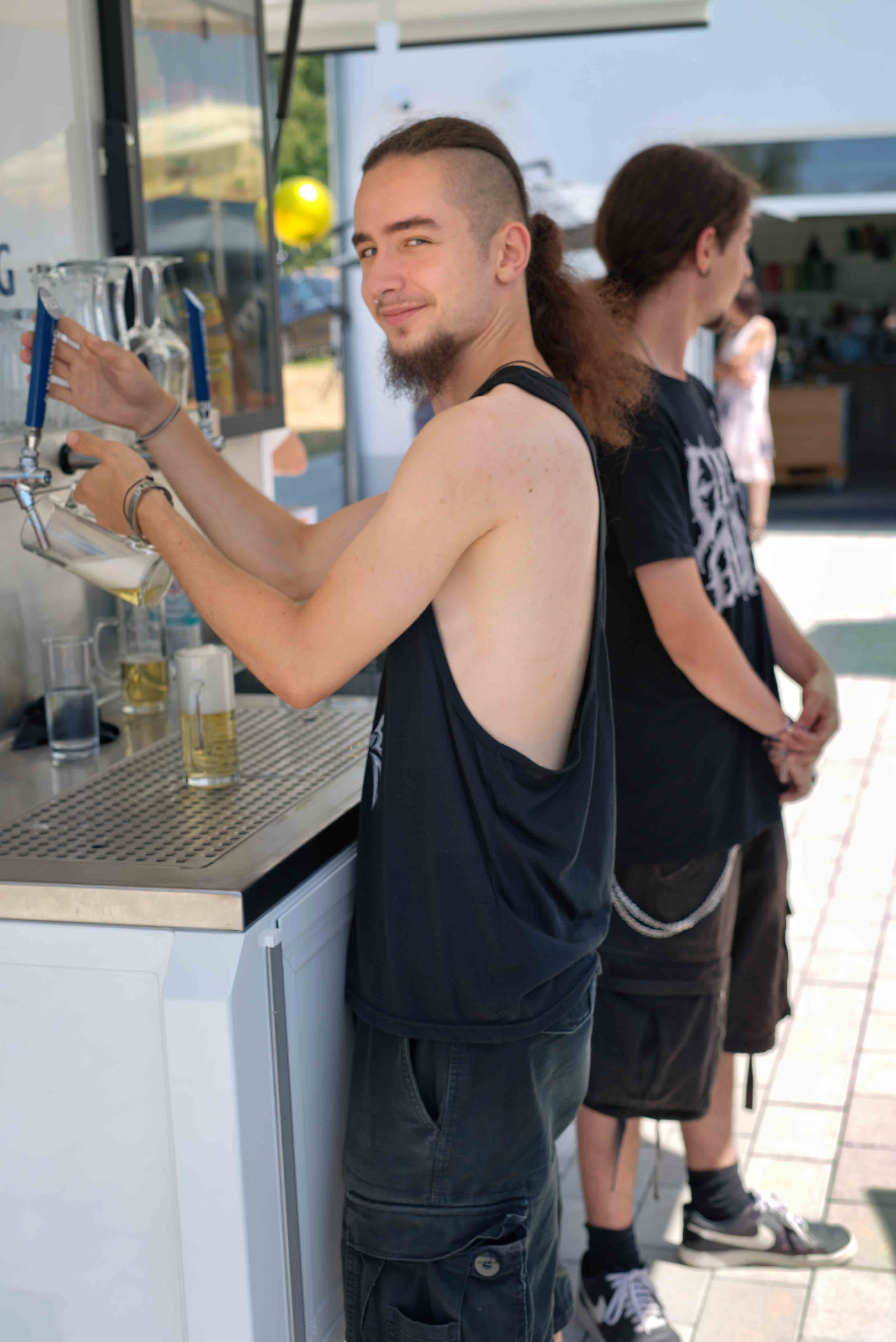 Young man with a beard and ponytail pouring a drink from a tap at an outdoor bar, smiling at the camera with another person standing nearby.