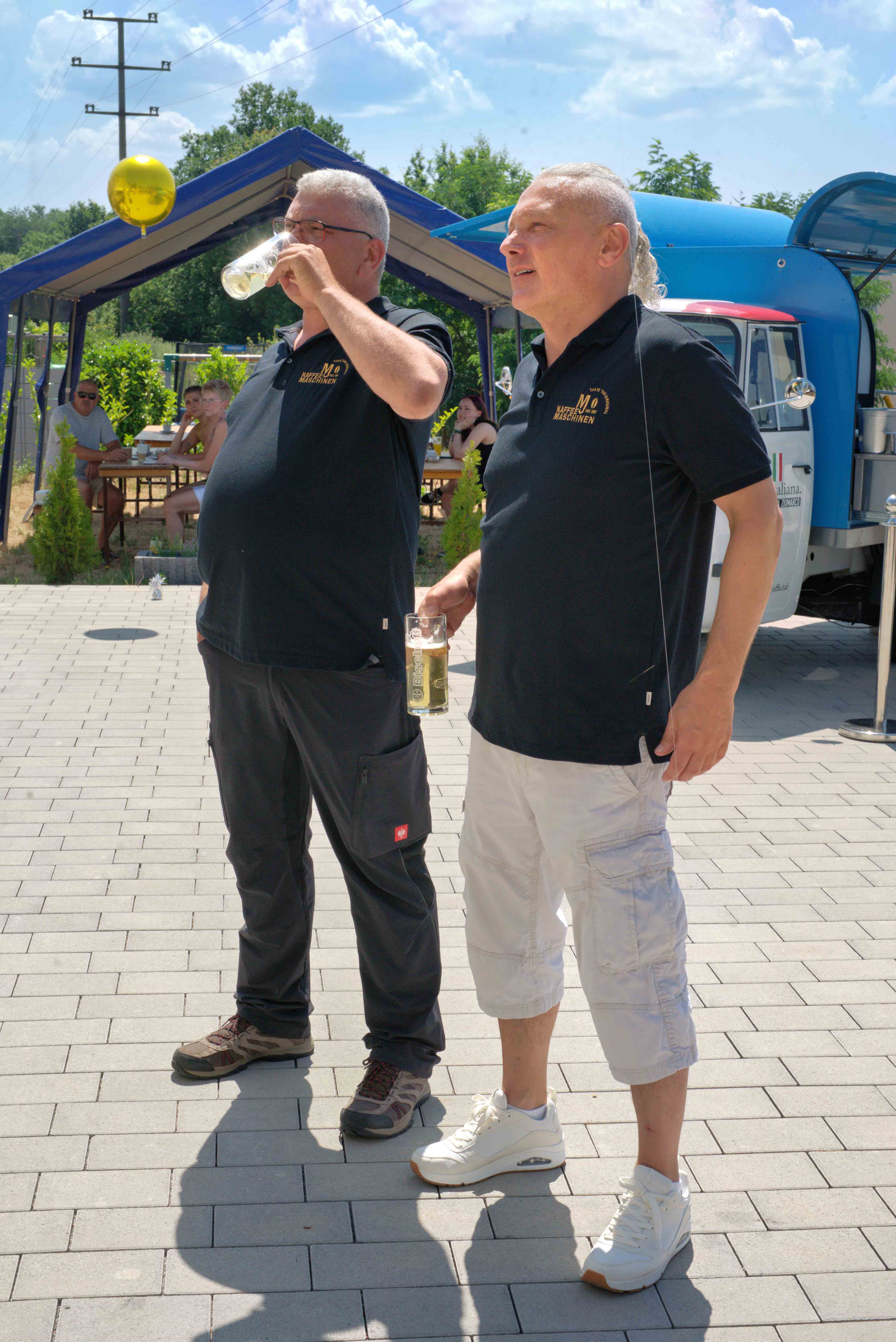 Two men in black polo shirts with logos stand outdoors on a sunny day, each holding a glass of beer.