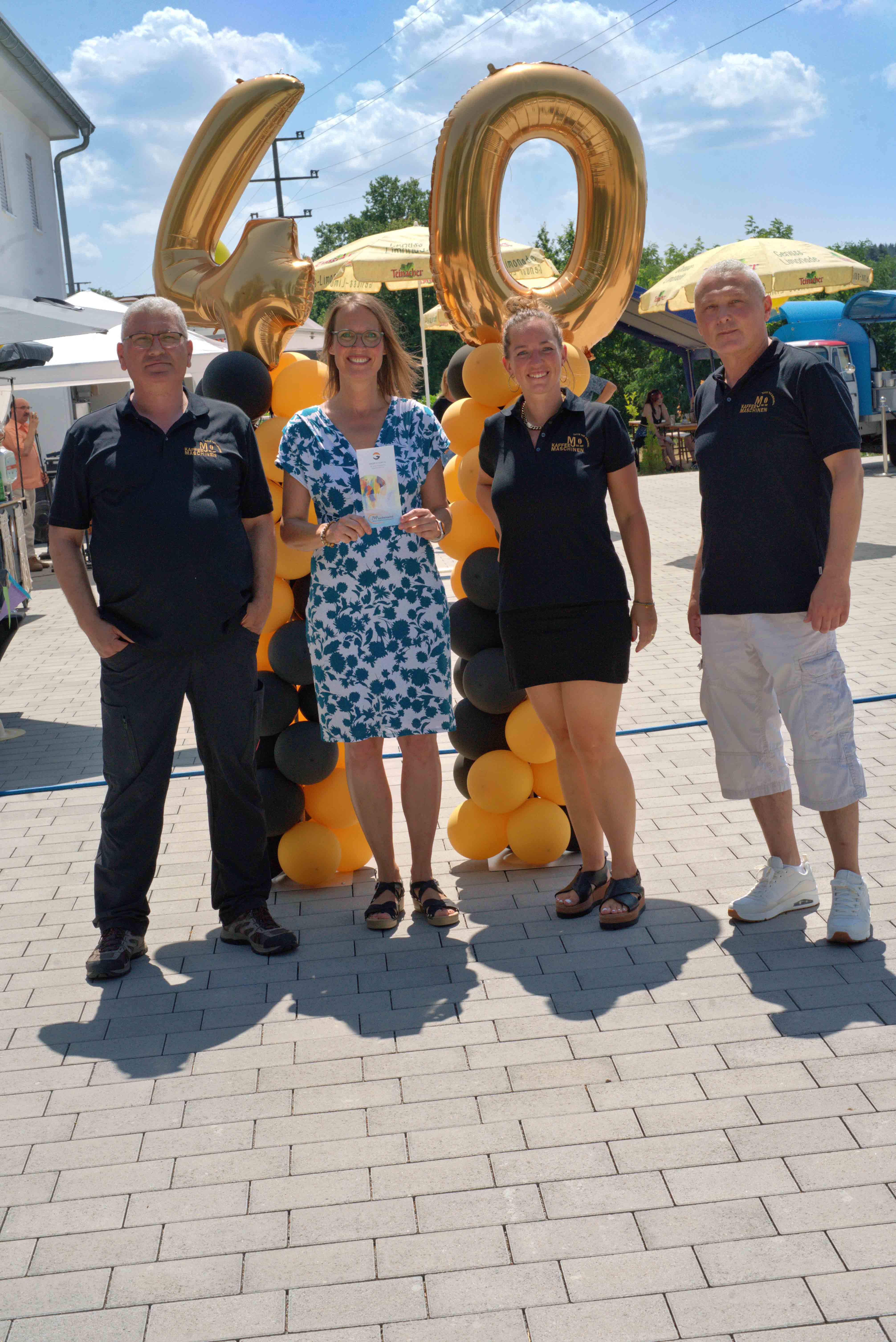 Four people standing outdoors on a sunny day in front of large gold balloon numbers 4 and 0 with black and orange balloon columns.