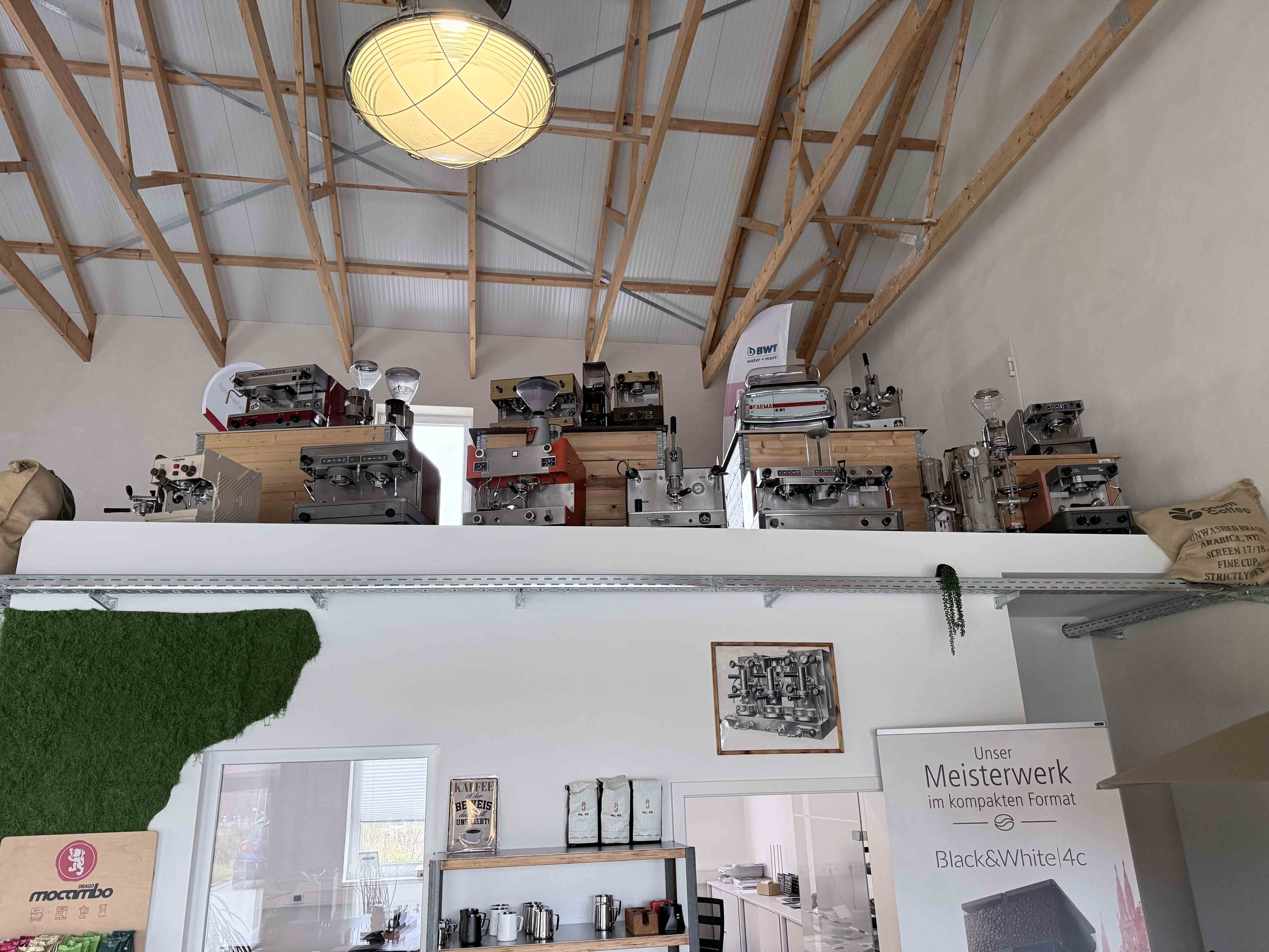 Interior of a room with wooden beams on the ceiling and a shelf displaying multiple vintage espresso machines and coffee grinders above a white wall with coffee-related decor and a large light fixture.