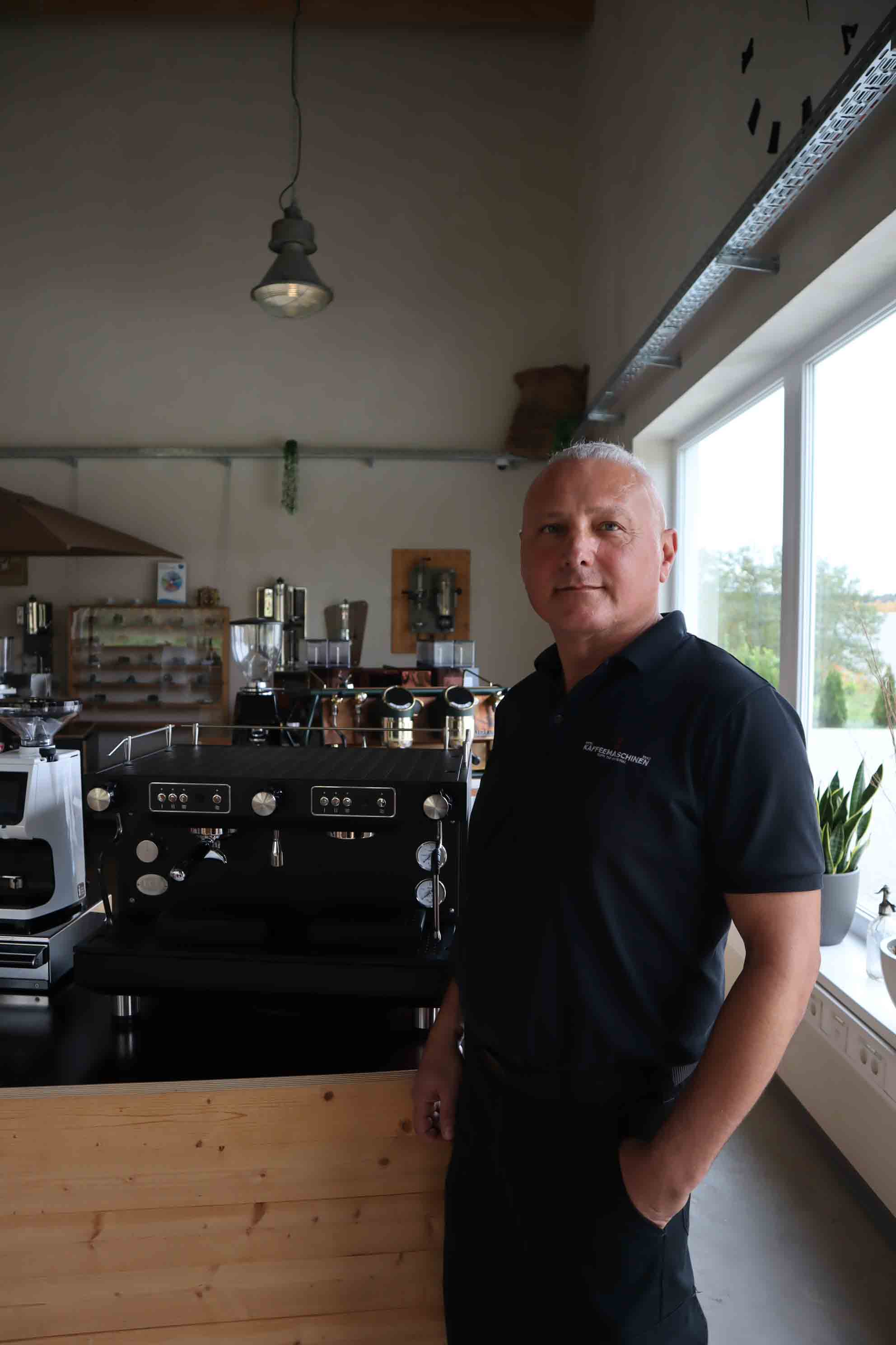Man in black polo shirt standing next to a large black coffee machine in a bright room with wooden counters and a large window.