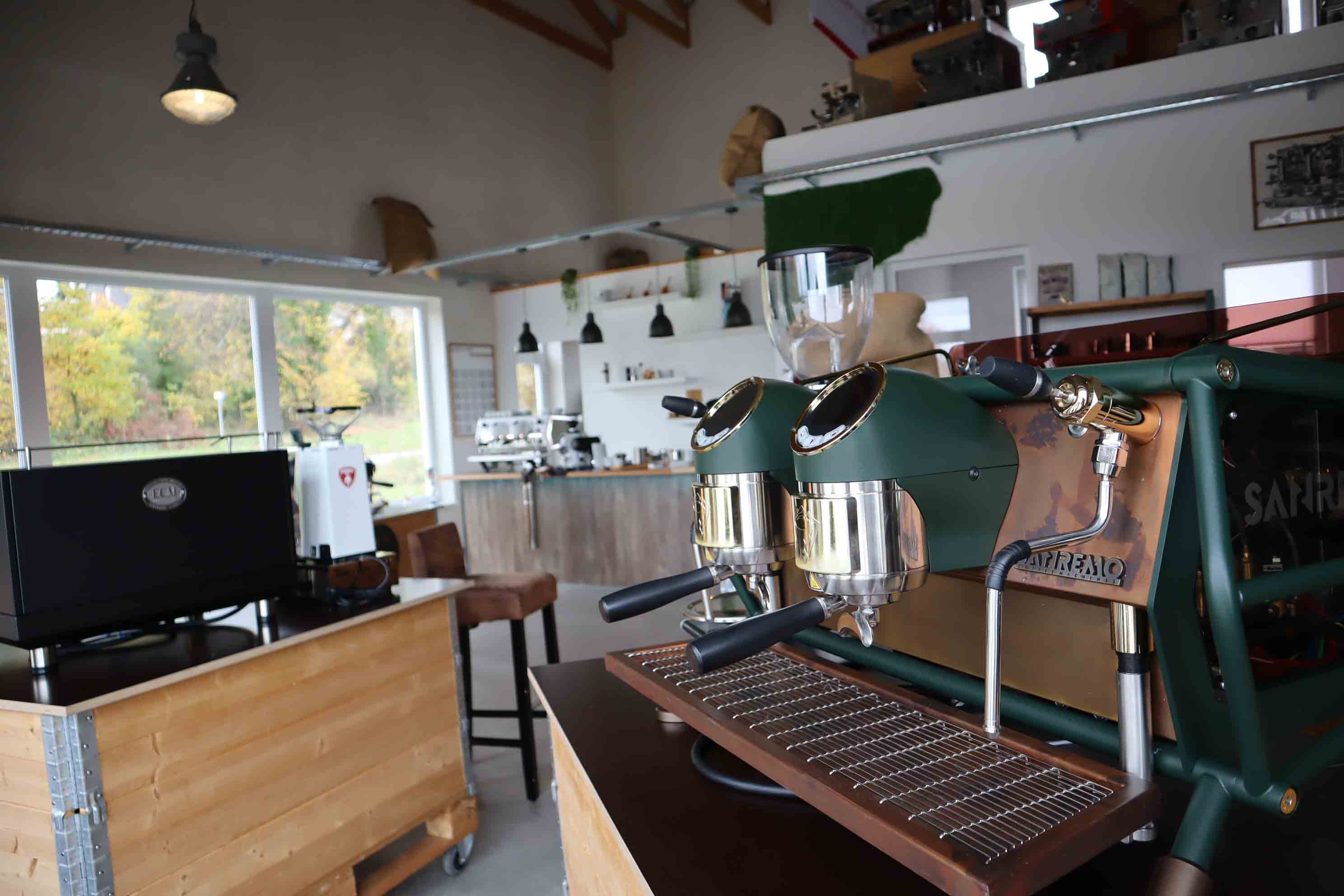 Modern coffee shop interior with a green dual-group espresso machine in the foreground and wooden counters with large windows showing autumn trees.