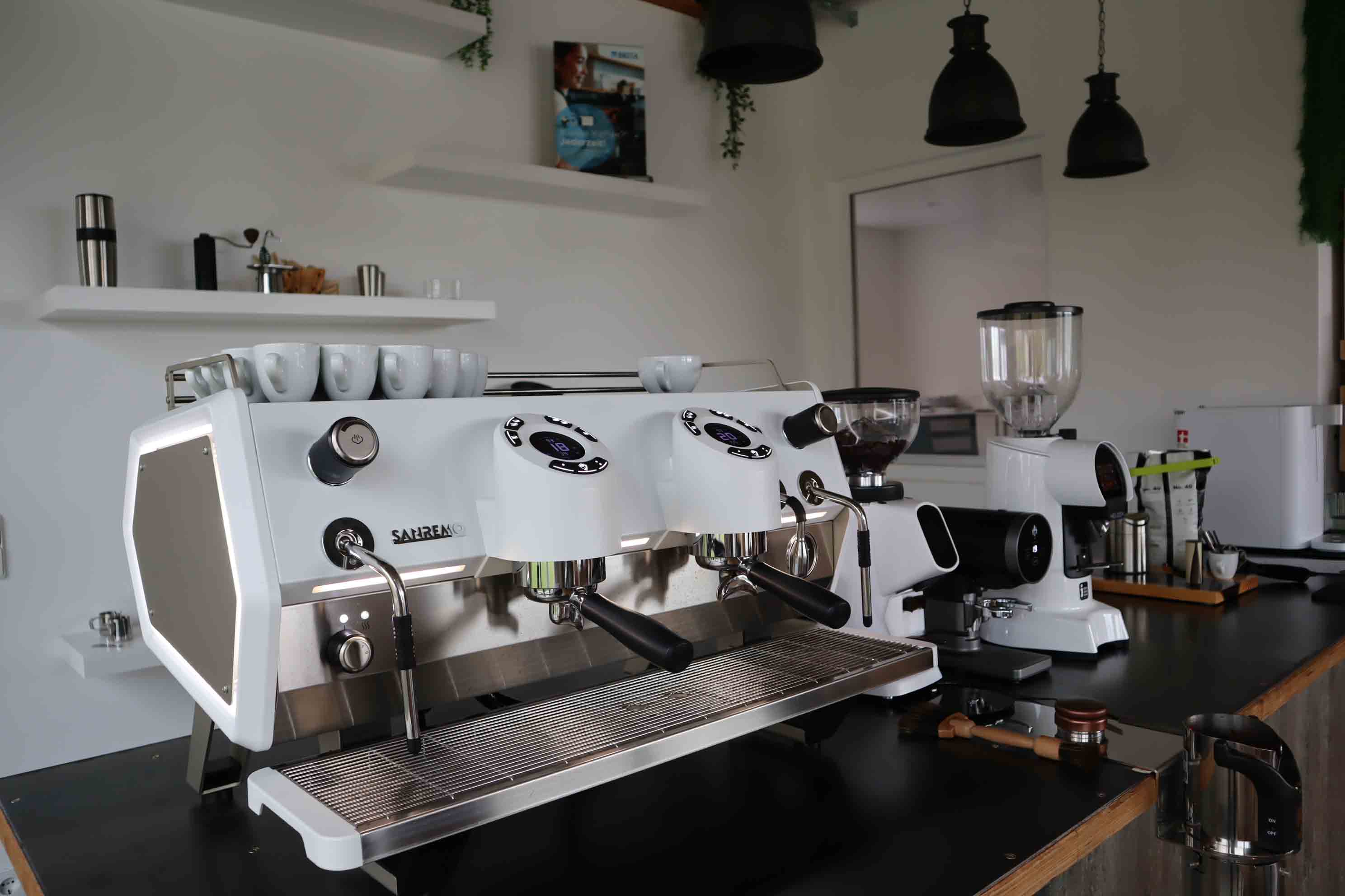 Modern white Sanremo espresso machine on a black counter with coffee grinder and cups in a café setting.