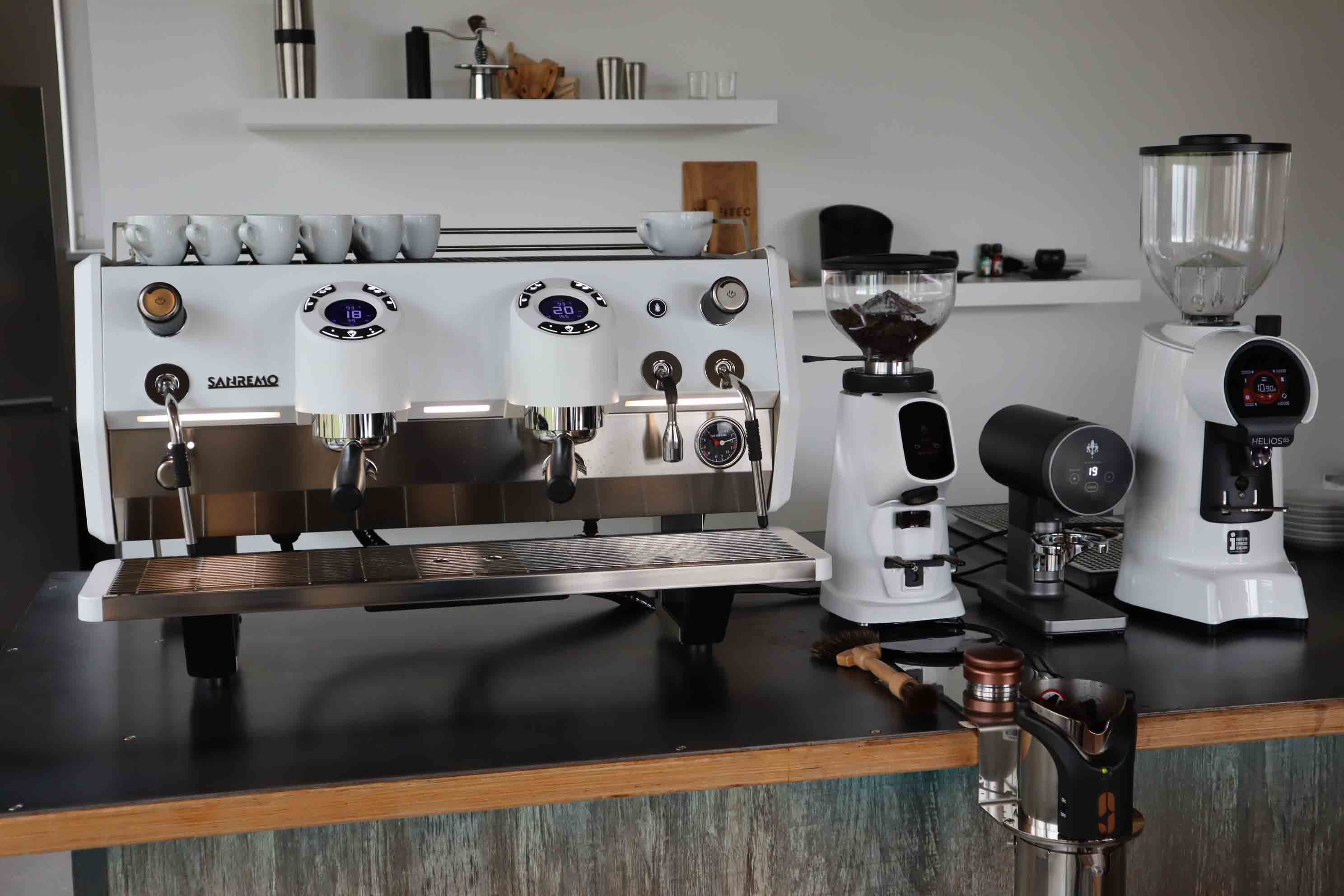 Commercial espresso machine with six white cups on top, accompanied by two coffee grinders and a tamper on a black countertop.