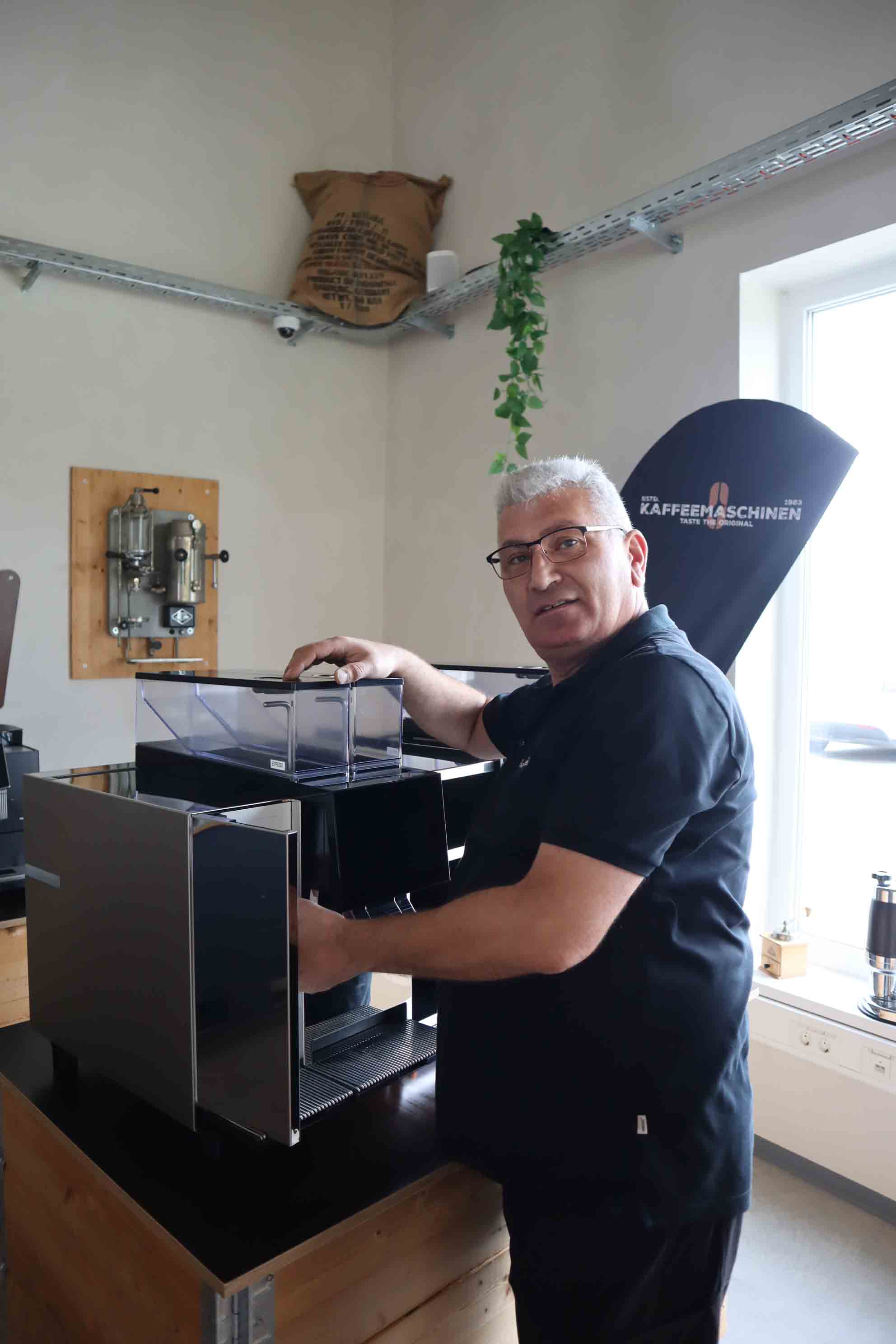 Man with glasses operating a modern black coffee machine in a bright room with a window and 'Kaffeemaschinen' banner.