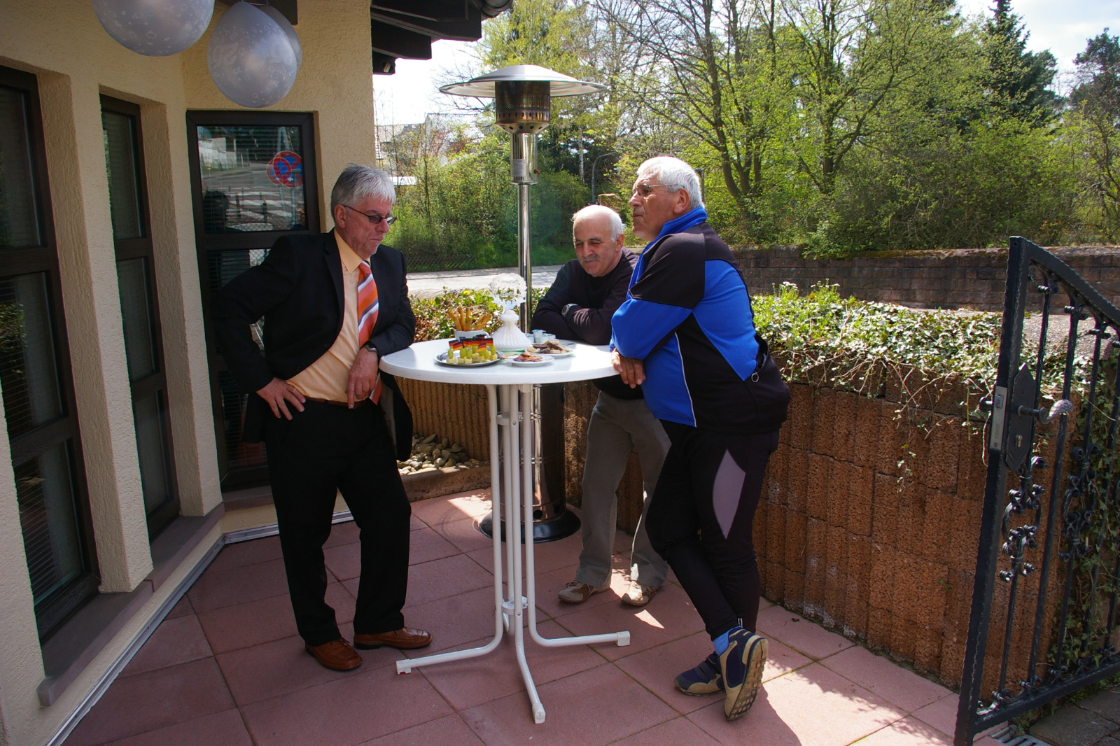 Three older men standing around a tall round outdoor table with snacks on a patio near a heater and greenery.