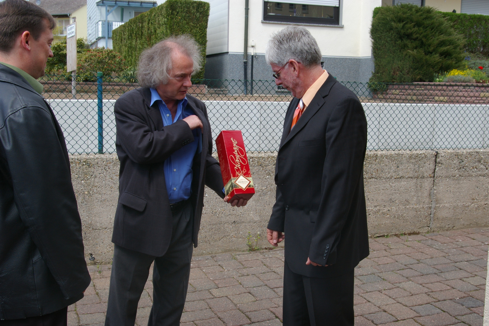 Three men dressed in suits and jackets standing outdoors, one man with gray hair holding a red gift box and showing it to the others.