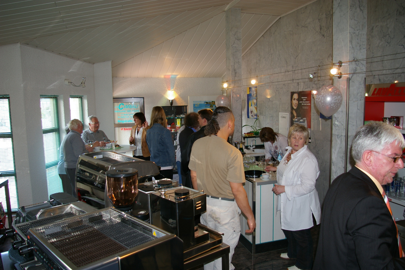 Group of people socializing inside a coffee shop near an espresso machine and counter with beverages.