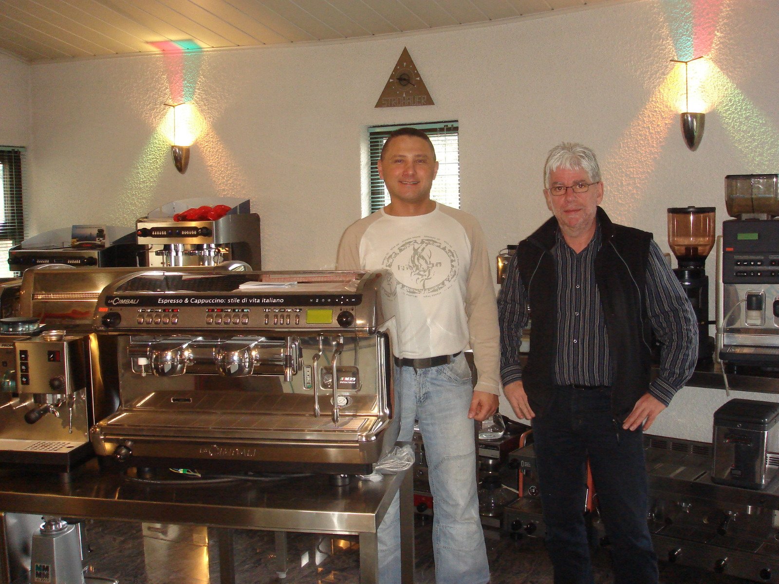 Two men standing inside a cafe or coffee shop with multiple espresso machines and coffee equipment on stainless steel tables.