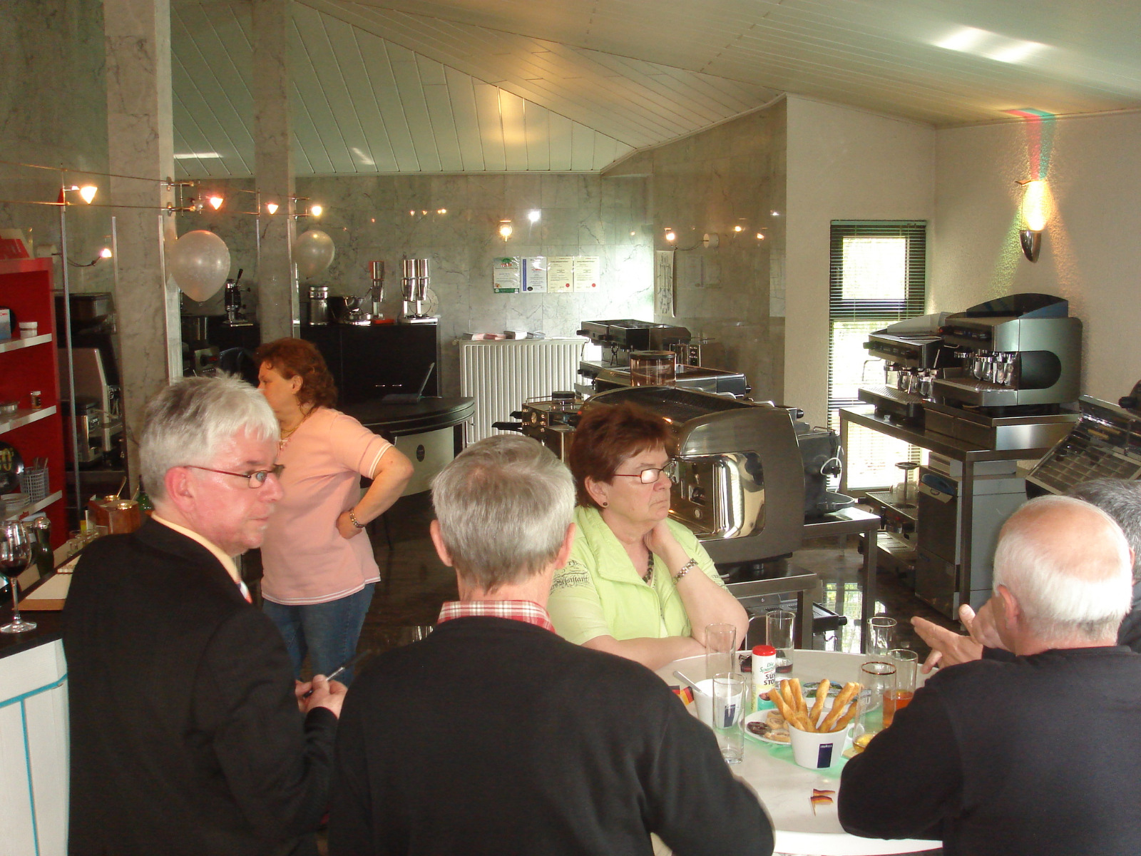 Group of adults gathered around a table in a café with coffee machines and snacks visible.